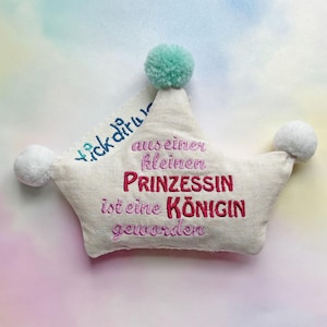 May include: A white crown with pink and blue embroidery that reads "tick diru aus einer kleinen Prinzessin ist eine Königin geworden". The crown has three white pom-poms.