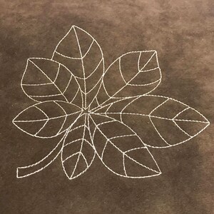 May include: A white embroidered design of a five-leafed clover on a brown fabric background.