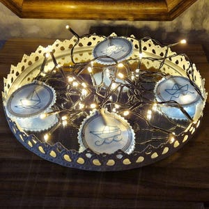 Peut inclure: Cinq ornements en tissu blanc brodés de fils bleus représentant différents bateaux sur un plateau blanc avec une surface miroir argentée. Le plateau est décoré d'une bordure en dentelle blanche et est entouré de guirlandes lumineuses blanches.