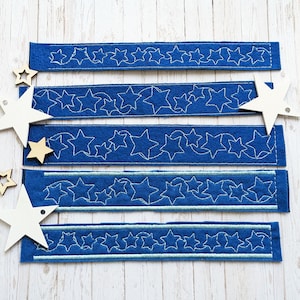 May include: Five blue felt strips with white embroidered star designs. The strips are arranged vertically on a white wooden surface.