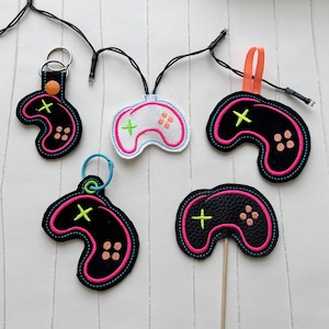 May include: A collection of embroidered game controller-shaped items in black, white, and pink. Some have keychains or ribbons attached. The controllers feature a cross, buttons, and are outlined in bright pink. A string of lights is in the background.