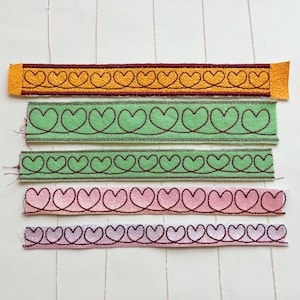 May include: Five different fabric trims with a heart pattern. The trims are in various colors, including yellow, green, pink, and white. The hearts are outlined in black thread.