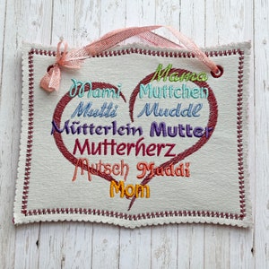 Könnte beinhalten: Ein weißes Stoffherz-Schild mit roten Nähten an den Rändern. Das Schild hat ein rotes Herz in der Mitte mit den Wörtern "Mama", "Mutchen", "Muddel", "Mütterlein", "Mutter", "Mutterherz", "Autsch Muddi" und "Mom", die in verschiedenen Farben um das Herz herum gestickt sind.
