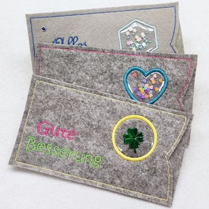 May include: Three gray felt envelopes with different embroidered designs. The top envelope has a blue and white design with the text "Alles Gute". The middle envelope has a pink heart with a clear window filled with colorful confetti. The bottom envelope has a green four-leaf clover in a yellow circle with the text "Gute Besserung" in pink and green.