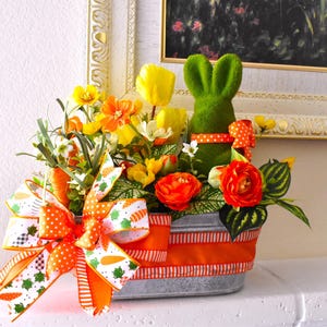 May include: A spring-themed floral arrangement in a galvanized metal container with a bright orange and white striped ribbon. The arrangement features yellow and orange flowers, green foliage, and a green felt bunny.