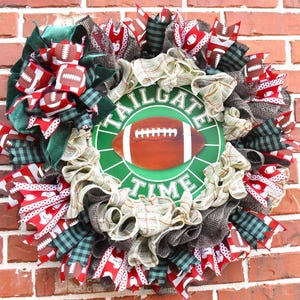 May include: A decorative wreath with a football theme. The wreath features a central green circle with a football graphic and the words "TAILGATE TIME." It is adorned with red, white, green, and brown ribbons and accents, perfect for game day.