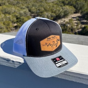 Può includere: Un cappello da camionista nero e grigio con una rete blu chiaro sul retro. Il cappello presenta una toppa in pelle marrone con il testo "Lake Travis 30.4300° N 97.9807° W" e una mappa del lago.