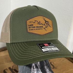 May include: A green and tan trucker hat with a brown patch that says "LAKE OF THE OZARKS 38.1380° N. 92.8104° W" and a map of the lake. The hat is made by Richardson and is adjustable.