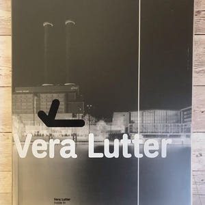 Puede incluir: Portada de libro con una fotografía en blanco y negro de un paisaje industrial. El título "Vera Lutter" está en letras blancas grandes, con el subtítulo "Inside In" debajo. Una flecha negra apunta hacia la izquierda.