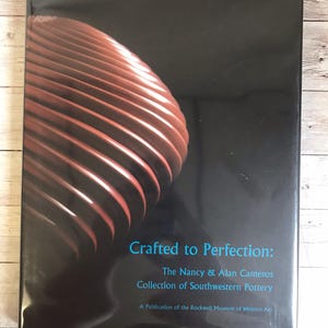 May include: A book cover with a dark background and a close-up of a brown, spiraling pottery design. The title "Crafted to Perfection" is in blue text, with the subtitle "The Nancy & Alan Cameros Collection of Southwestern Pottery."