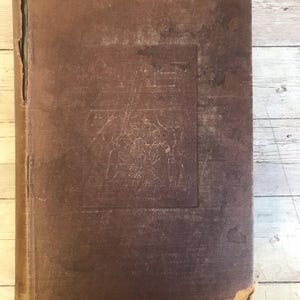 May include: A vintage book with a worn, brown cover. The cover has a textured appearance with a rectangular, slightly darker area in the center. The edges show signs of wear and tear, suggesting age and use. The book's spine is visible on the left side.