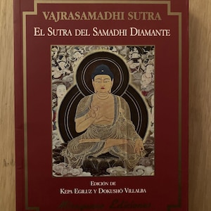 May include: A red book cover with gold lettering that reads "VAJRASAMADHI SUTRA" and "EL SUTRA DEL SAMADHI DIAMANTE". The cover features a painting of a seated Buddha with a golden halo and a lotus flower.