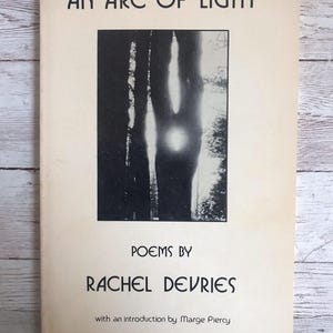 May include: A vintage book cover with the title "AN ARC OF LIGHT" in black capital letters. Below the title is a black and white image of trees with a bright light. The author's name, RACHEL DEVRIES, is printed below the image.