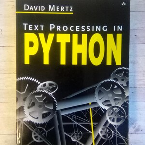 May include: A book titled "Text Processing in Python" by David Mertz. The cover is black with yellow text and a graphic of gears and mechanical structures in shades of gray. The book is on a light wood grain surface.