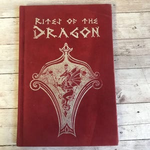 May include: A red hardcover book with the title "Rites of the Dragon" in silver lettering. The cover features a silver embossed design of a dragon within a decorative frame.