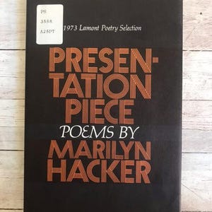 May include: A vintage book titled "Presentation Piece Poems by Marilyn Hacker" with orange text on a black cover. The book is a 1973 Lamont Poetry Selection. A white sticker with text is in the upper left corner.