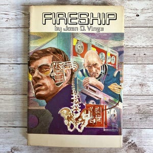 May include: A vintage paperback book titled "FIRESHIP" by Joan D. Vinge. The cover art features a futuristic illustration with two men, one with a partially exposed mechanical face, and a complex background.