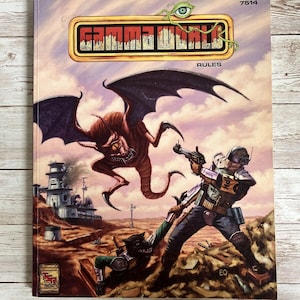 May include: A vintage paperback book cover for the Gamma World role-playing game. The cover features a colorful illustration of a futuristic scene with a winged creature, a human character with a gun, and a desert landscape. The title "Gamma World" is printed in bold letters on a yellow banner.