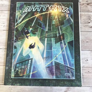 May include: A black and green book cover with the title "MATRIX" in white letters. The cover features a digital art illustration of a person in a white suit standing in a futuristic cityscape with green and blue screens.
