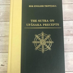 May include: A green hardcover book with a gold spine and title. The title is "The Sutra on Upasaka Precepts" and the book is part of the BDK English Tripitaka series. A gold Dharmachakra symbol is on the front cover.