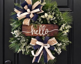 May include: A circular wreath made of green pine needles and frosted foliage, featuring two decorative bows in navy blue, white, and tan patterned ribbon. A rectangular wooden sign with a dark brown finish displays the word hello in white script.