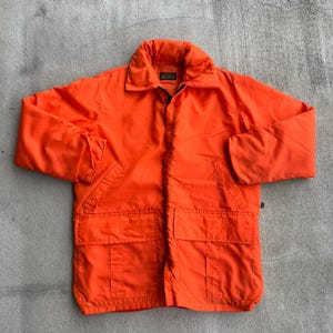 May include: Bright orange jacket with a large collar, two front pockets, and a zipper closure. The jacket has a quilted appearance and a small label near the collar. The jacket is made of a lightweight material.