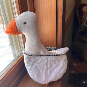 May include: A white goose-shaped fabric basket with a black and white checkered interior. The goose has a white body, an orange beak, and a black eye. The basket is quilted with a diamond pattern.