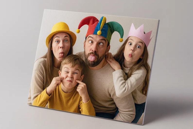 Op de afbeelding: Een canvas print met een gezin van vier dat gekke gezichten trekt. De volwassenen dragen een gele hoed en een narrenmuts, terwijl de kinderen een geel shirt en een roze kroon dragen. De afbeelding is schuin.