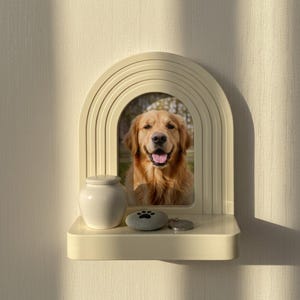 May include: A cream-colored pet memorial shelf with an arched frame holding a photo of a golden retriever. A small white urn, a stone with a paw print, and a metal tag sit on the shelf. The shelf is mounted on a textured wall.