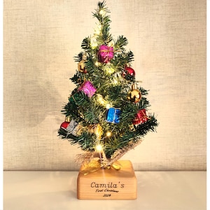 Puede incluir: Un pequeño árbol de Navidad decorado con luces y adornos. El árbol está adornado con adornos coloridos en forma de regalo y bolas doradas. La base de madera tiene la inscripción "Camila's First Christmas 2024".