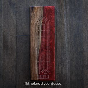 Peut inclure: Un jeu de cribbage en bois avec un design bicolore. Un côté est en bois brun clair, l'autre en résine rouge foncé. Le jeu comporte des lignes et des trous gravés pour les chevilles, avec le texte "@theknottycontessa" en bas.