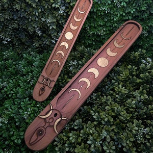 May include: Two wooden incense holders with a crescent moon design. The holders are brown with gold accents and have a triple moon symbol at the bottom.