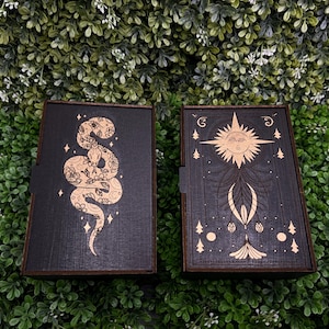 May include: Two wooden boxes with intricate laser-cut designs. The box on the left features a snake with floral details, while the box on the right depicts a sun with a face and radiating rays.