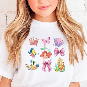 A young girl with long blonde hair wearing a white t-shirt with a colorful graphic design featuring characters from the Disney movie "The Little Mermaid".