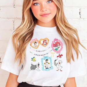 A young girl with long blonde hair wearing a white t-shirt with a graphic of Disney characters and a black skirt.
