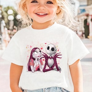 A young girl with blonde hair is smiling and wearing a white t-shirt with a graphic of two cartoon characters, Jack Skellington and Sally.