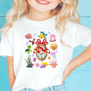 A young girl with blonde hair is smiling and wearing a white t-shirt with a colorful graphic of underwater sea creatures.