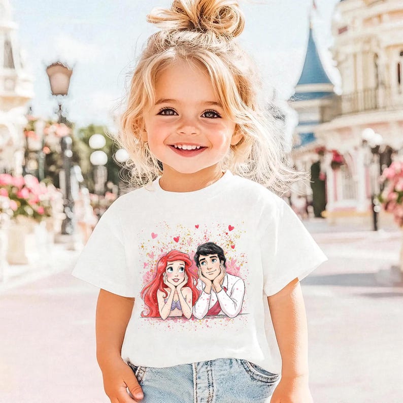 A young girl with blonde hair is smiling and wearing a white t-shirt with a Disney character graphic.