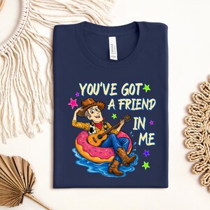 A blue t-shirt with a cartoon image of a man playing a guitar while sitting on a donut, surrounded by stars and text that says "You've Got a Friend in Me".