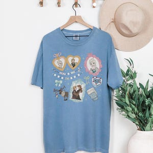 A blue t-shirt with various cartoon-style illustrations and characters, including a couple, a cup, and other decorative elements, hanging on a wooden hanger.