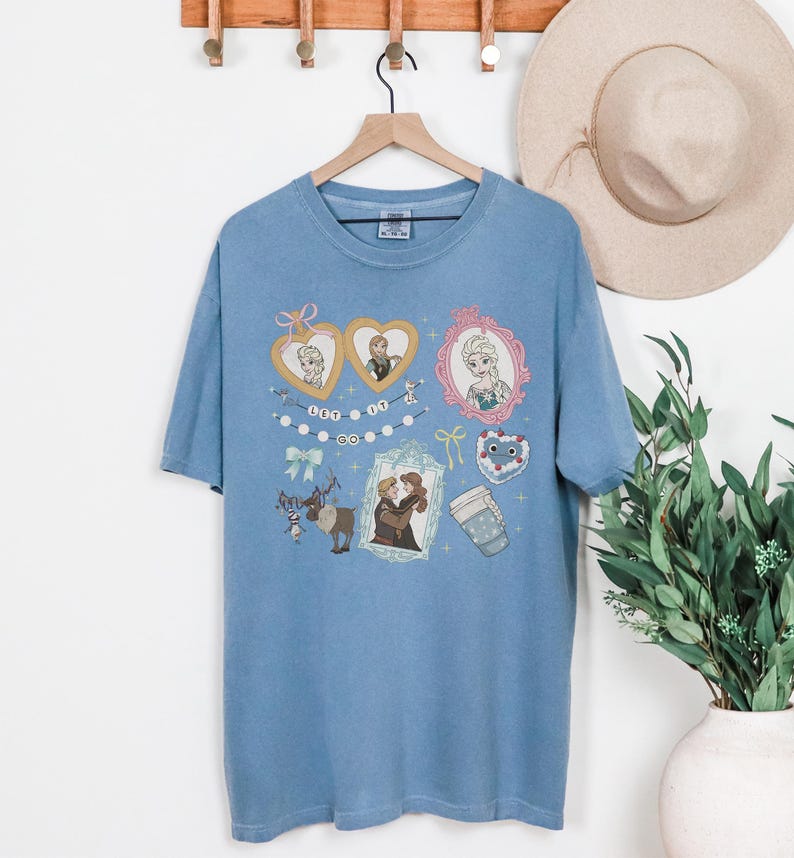 A blue t-shirt with various cartoon-style illustrations and characters, including a couple, a cup, and other decorative elements, hanging on a wooden hanger.