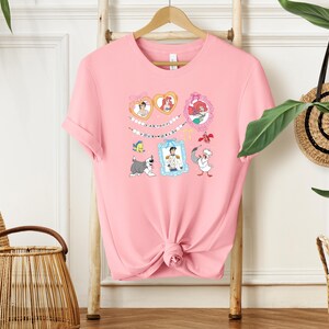 A pink t-shirt with a cartoon character design hangs on a wooden hanger, surrounded by various decorative items such as a woven basket and a potted plant.