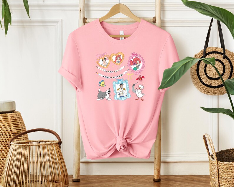 A pink t-shirt with a cartoon character design hangs on a wooden hanger, surrounded by various decorative items such as a woven basket and a potted plant.