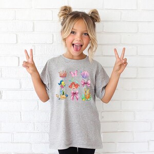 A young girl with blonde hair is making a peace sign with her hands, wearing a gray t-shirt with a colorful graphic of mermaids and other sea creatures.