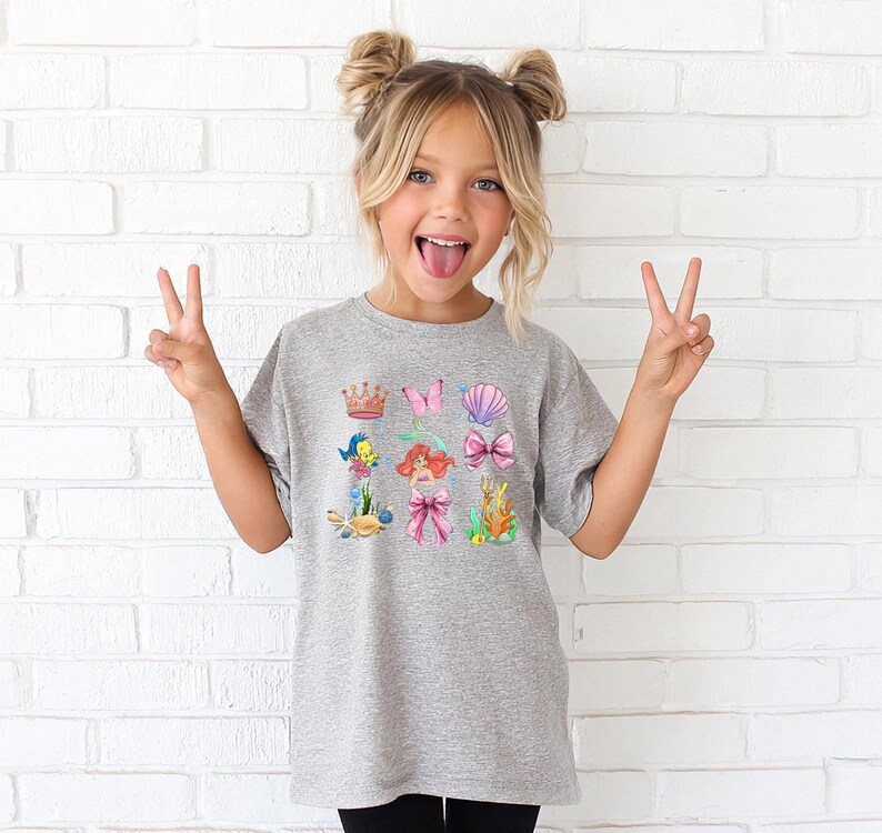 A young girl with blonde hair is making a peace sign with her hands, wearing a gray t-shirt with a colorful graphic of mermaids and other sea creatures.