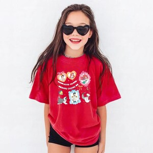 A young girl wearing a red t-shirt with cartoon characters and smiling at the camera.