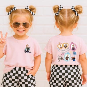 A young girl with blonde hair and a pink shirt with a cartoon character on it is smiling and making a peace sign.