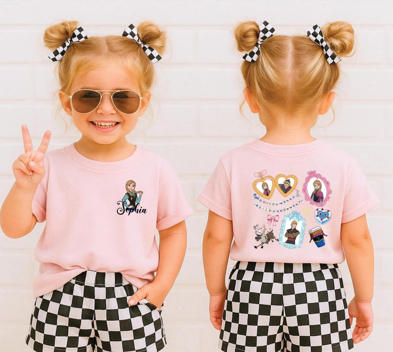 A young girl with blonde hair and a pink shirt with a cartoon character on it is smiling and making a peace sign.