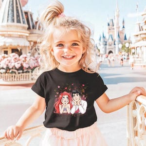 A young girl with blonde hair is smiling and posing in front of a Disney theme park, wearing a black t-shirt with a Disney character design.