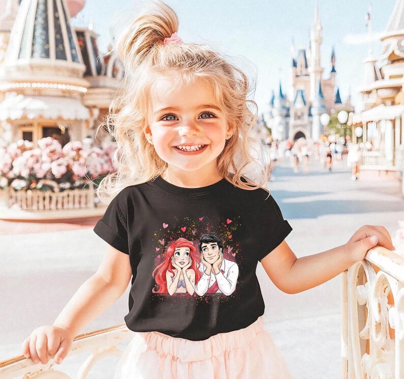 A young girl with blonde hair is smiling and posing in front of a Disney theme park, wearing a black t-shirt with a Disney character design.